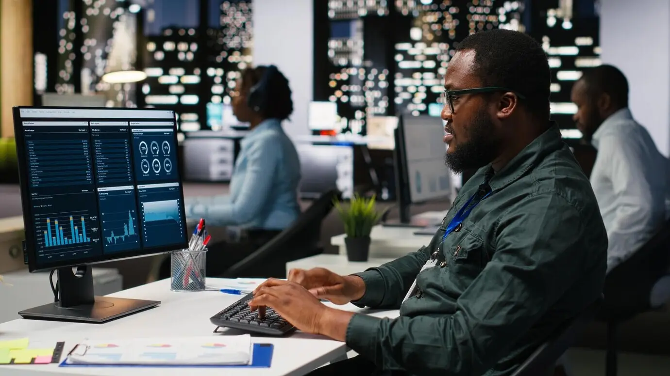 Empleado de oficina revisando métricas de KPI en el monitor y analíticas en el escritorio, construyendo una visión y